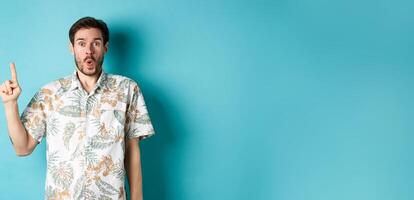 Surprised caucasian tourist in hawaiian shirt, pointing finger up and say wow, checking out promotion, standing on blue background photo