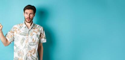 Handsome guy wearing summer shirt on vacation, pointing and looking aside at empty space, making amazed face, standing on blue background photo