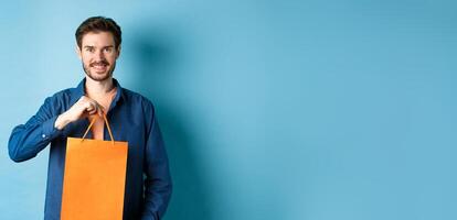 Handsome young man with beard, smiling and showing shopping bag, buying something in store, standing on blue background photo