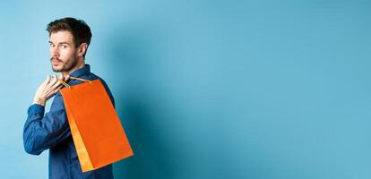 Rear view of happy man holding shopping back on shoulder and looking behind, standing on blue background photo
