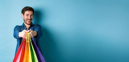 Cheerful young guy stretch out hands with shopping bags and smiling, giving you gifts, standing on blue background photo