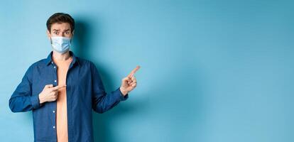 Covid and healthcare concept. Worried young man in medical mask raising eyebrow, pointing finger right with doubtful and nervous face, standing on blue background photo