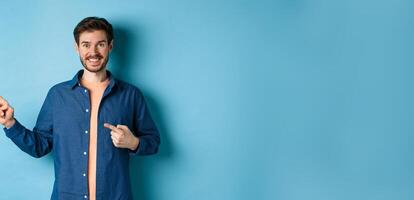 Happy smiling man pointing fingers left at logo, showing advertisement, standing on blue background photo