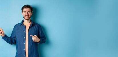 Happy smiling caucasian man showing advertisement, pointing fingers left at empty space, standing on blue background photo