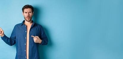 Shocked and offended young man frowning and looking hurt, pointing fingers left at empty space, complaining on something, standing on blue background photo