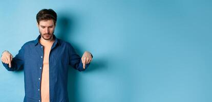 Image of modern guy in casual clothes, looking and pointing fingers down at empty space, reading logo, standing on blue background photo