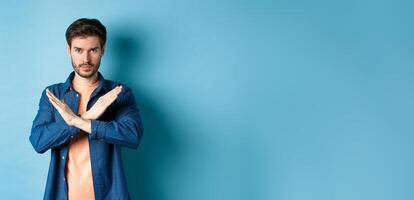 Serious caucasian man tell no, showing cross stop gesture and looking confident at camera, prohibit something bad, standing on blue background photo