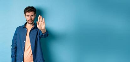 Upset man frowning and asking to stop, stretch out hand to prohibit or disagree with something bad, standing on blue background photo