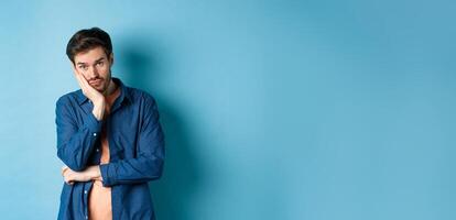 Sad and bored man lean face on hand, looking at camera empty-headed, standing bothered on blue background photo