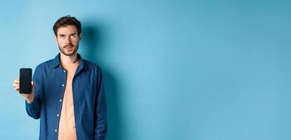 Annoyed young guy showing empty smartphone screen and roll eyes up bothered, standing on blue background photo