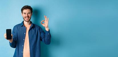 Handsome young man showing okay gesture and empty smartphone screen, recommending app or company, standing on blue background photo