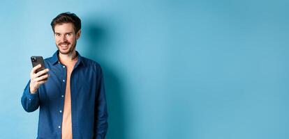 Attractive caucasian man using mobile phone, looking happy and smiling at camera, standing on blue background photo