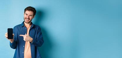 Smiling caucasian man showing on empty smartphone screen, pointing at mobile phone and looking satisfied, standing on blue background photo