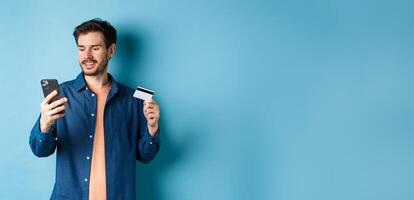 E-commerce concept. Image of handsome caucasian man paying online, looking at mobile phone screen and holding plastic credit card, blue background photo