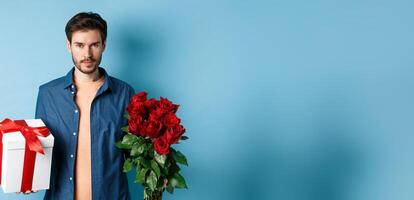 Love and Valentine day concept. Passionate man looking confident at camera, holding gift box and red roses for romantic date, standing over blue background photo