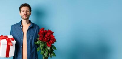 Love and Valentines day concept. Romantic boyfriend pucker lips for kiss, bring bouquet of red roses and gifts on date, standing over blue background photo