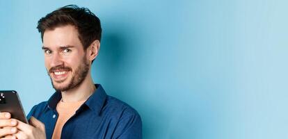 Close-up of cheerful bearded man using smartphone, smiling confident at camera, standing on blue background photo