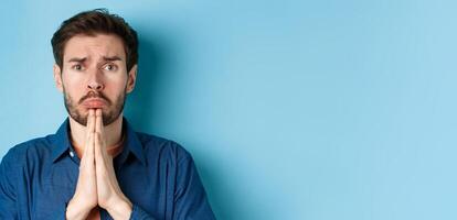 Sad young man begging for help, apoligizing and sobbing miserable, standing on blue background photo