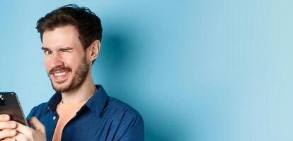 Portrait of handsome young man with beard, winking and smiling at camera, using smartphone, standing on blue background photo
