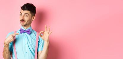 Image of young handsome guy in bow-tie and holiday outfit, pointing at himself and showing okay gesture, self-promoting on pink background photo