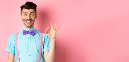 Handsome caucasian man with moustache smiling and showing okay sign, approve something good, praising excellent choice, standing amused on pink background photo