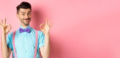 Portrait of handsome man with moustache and bow-tie, showing okay gestures and smiling in approval, recommending good offer, standing over pink background photo