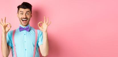 Amazed caucasian guy showing okay gestures and smiling, recommending cool thing, praising awesome offer, standing in pink background photo
