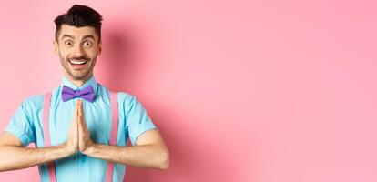 Image of smiling young man asking please, looking grateful at camera, Guy in bow-tie say thank you, standing over pink background photo