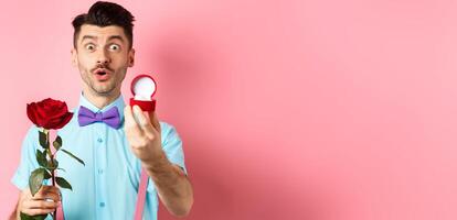 Valentines day. Funny man with moustache and bow-tie making proposal, showing engagement ring and propose with red rose, standing over pink background photo