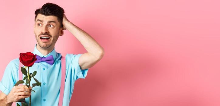 nervioso hombre esperando para su fecha en san valentin día, participación rojo Rosa y mirando confuso oblicuo, rascarse cabeza ansiosamente, en pie en rosado antecedentes foto