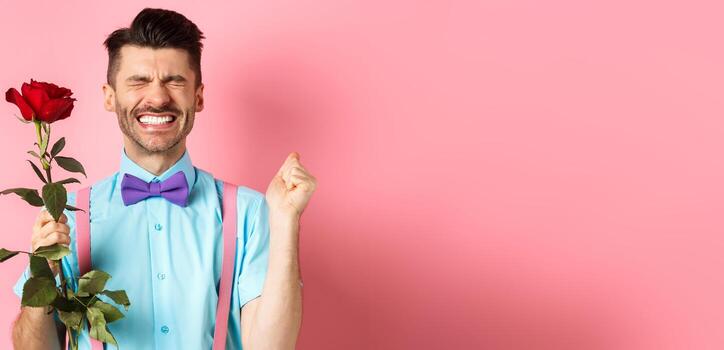 romance y san valentin día concepto. esperanzado hombre sensación emocionado antes de fecha, esperando para amante con rojo Rosa y Orando, en pie terminado rosado antecedentes foto