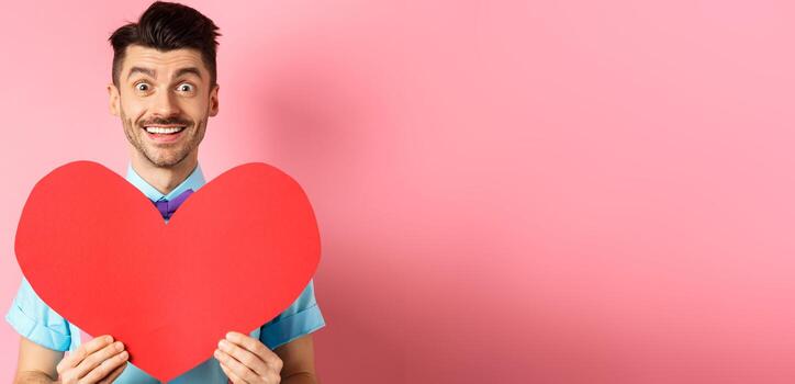 esperanzado hombre en amor demostración rojo corazón firmar, sonriente a cámara, esperando para alma gemela en san valentin día, en pie en rosado antecedentes foto