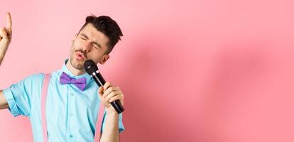 Party and festive events concept. Funny guy singing in microphone, raising finger up as reaching high note in song, standing on pink background photo