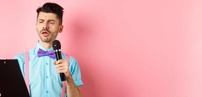 Holidays concept. Serious-looking man reading script from clipboard, holding microphone, entertain people on festive event, standing on pink background photo