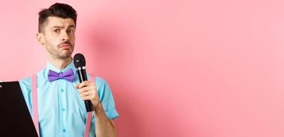 Holidays concept. Funny man with bow-tie making speech, perform on party events, holding script on clipboard and microphone, entertaining you, standing over pink background photo