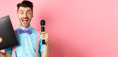 Holidays concept. Happy young man entertainer smiling at camera, holding clipboard and microphone, making speech on party event, standing over pink background photo