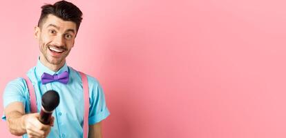 Smiling guy with moustache and bow-tie asking for comments or interview, stretch out hand with microphone, standing on pink background photo