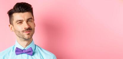 Close-up of smiling young guy with moustache, squinting intrigued, listening to something interesting, standing in bow-tie on pink background photo