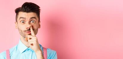Close-up of funny caucasian man picking nose, staring silly at camera, standing on pink background photo