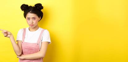 Sad and gloomy asian girl with glamour makeup, frowning unfair and pointing left at logo, looking disappointed, standing on yellow background photo