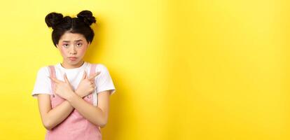 Indecisive timid asian girl pointing sideways, showing left and right ways and sulking upset, standing sad on yellow background photo