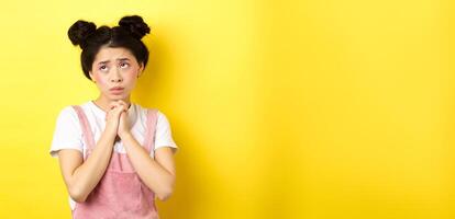 Sas asian girl holding hands in pray, pleading god, looking up and begging, standing on yellow background photo
