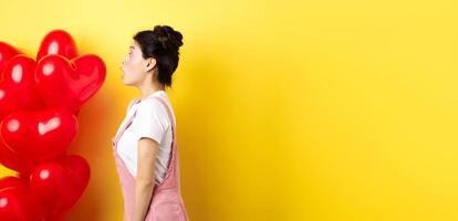 Valentines day and relationship concept. Profile of surprised asian girl staring left at logo, standing near heart balloons, yellow background photo