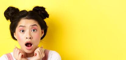 Head shot of shocked asian girl with hairbuns and glamour makeup, open mouth and looking startled at camera, standing against yellow background photo