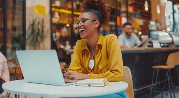 Young businesswoman with glasses sitting at a table in a coffee shop, writing notes and smiling while using a laptop computer during a work break photo