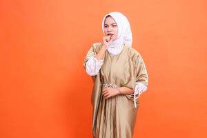 expression of a beautiful indonesia woman coughing candidly clenching her fist in front of her mouth wearing a kaftan Muslim dress with a hijab, isolated on an orange background. Concept of promotion photo