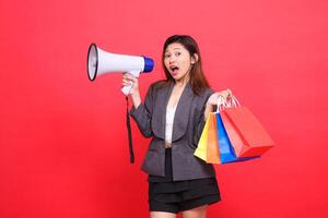 Asian sales woman shouts cheerfully at the camera through a megaphone speaker and holds up a shopping paper bag wearing a gray jacket on a red background. for transaction, lifestyle and business photo