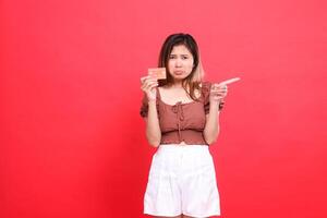 gesture of a sad, disappointed indonesia woman holding a debit credit card while pointing left, wearing a brown blouse on a red background. for transaction, technology and advertising concepts photo