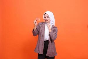 mature Asian woman in hijab suit frowning holding plastic debit credit card while hand in front of mouth up isolated on orange background. satisfied with service for financial, transaction, service photo