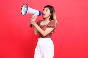 Elegant candid indonesia woman's expression shouting with both hands holding an audio megaphone wearing a brown blouse top and shorts on a red background. for broadcast, technology and advertising photo
